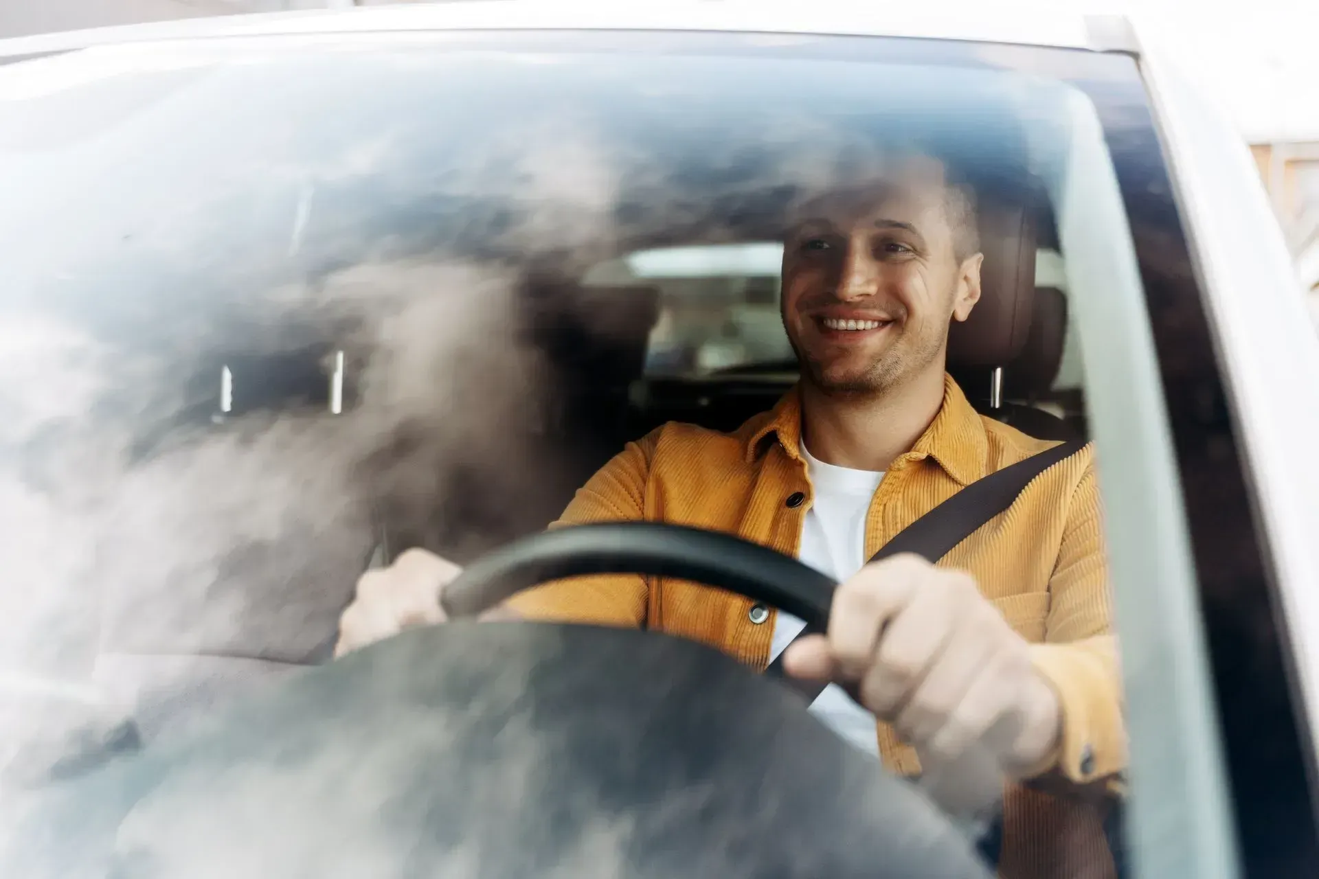 A smiling person wearing a mustard yellow shirt drives a car, hands on the steering wheel, seen through the windshield.