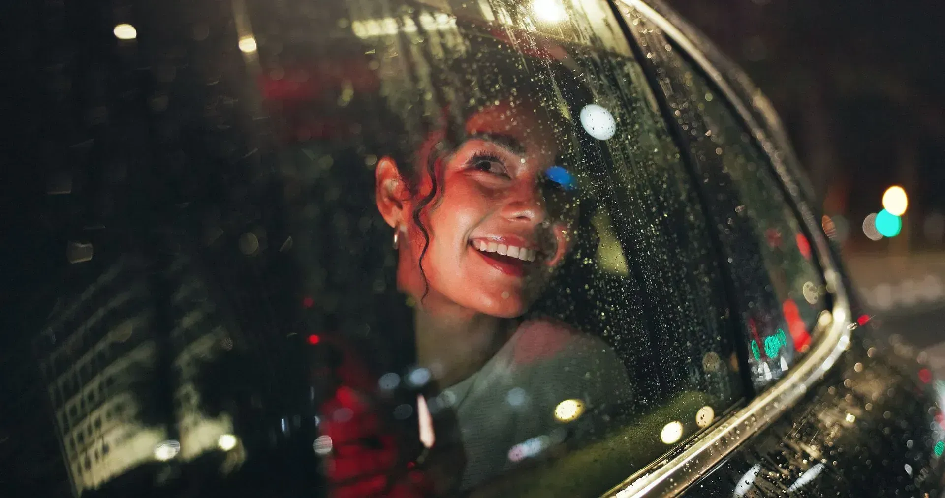 A person smiling while looking through a rain-streaked car window at night, with blurred city lights in the background.