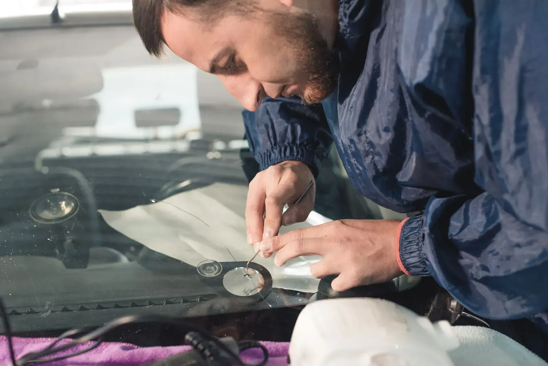 A person in a blue jacket carefully repairs a crack in a car windshield.