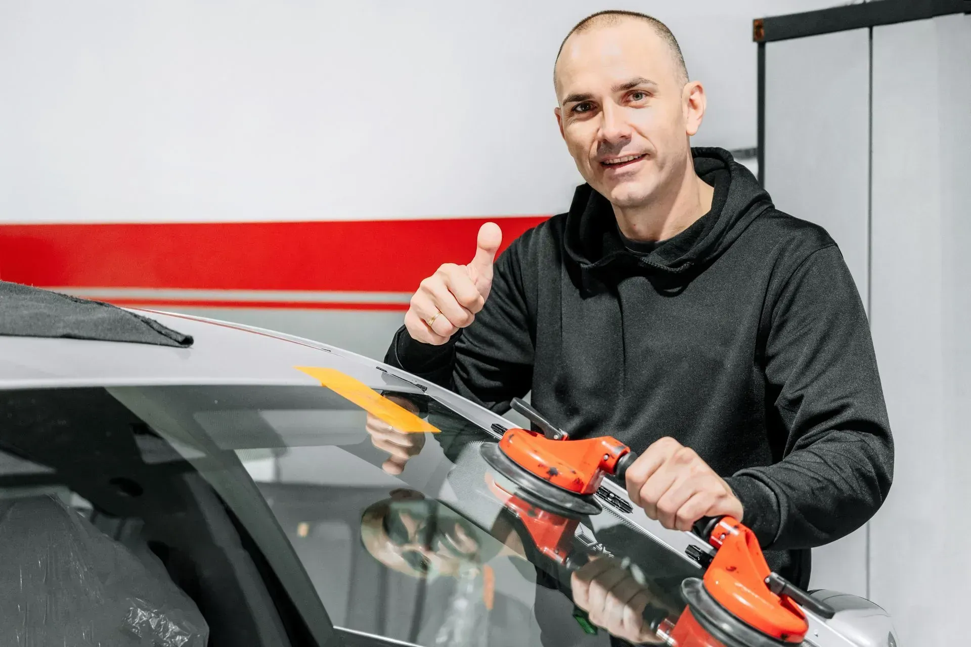 A technician giving a thumbs-up while using a suction tool to install a new car windshield in a garage.