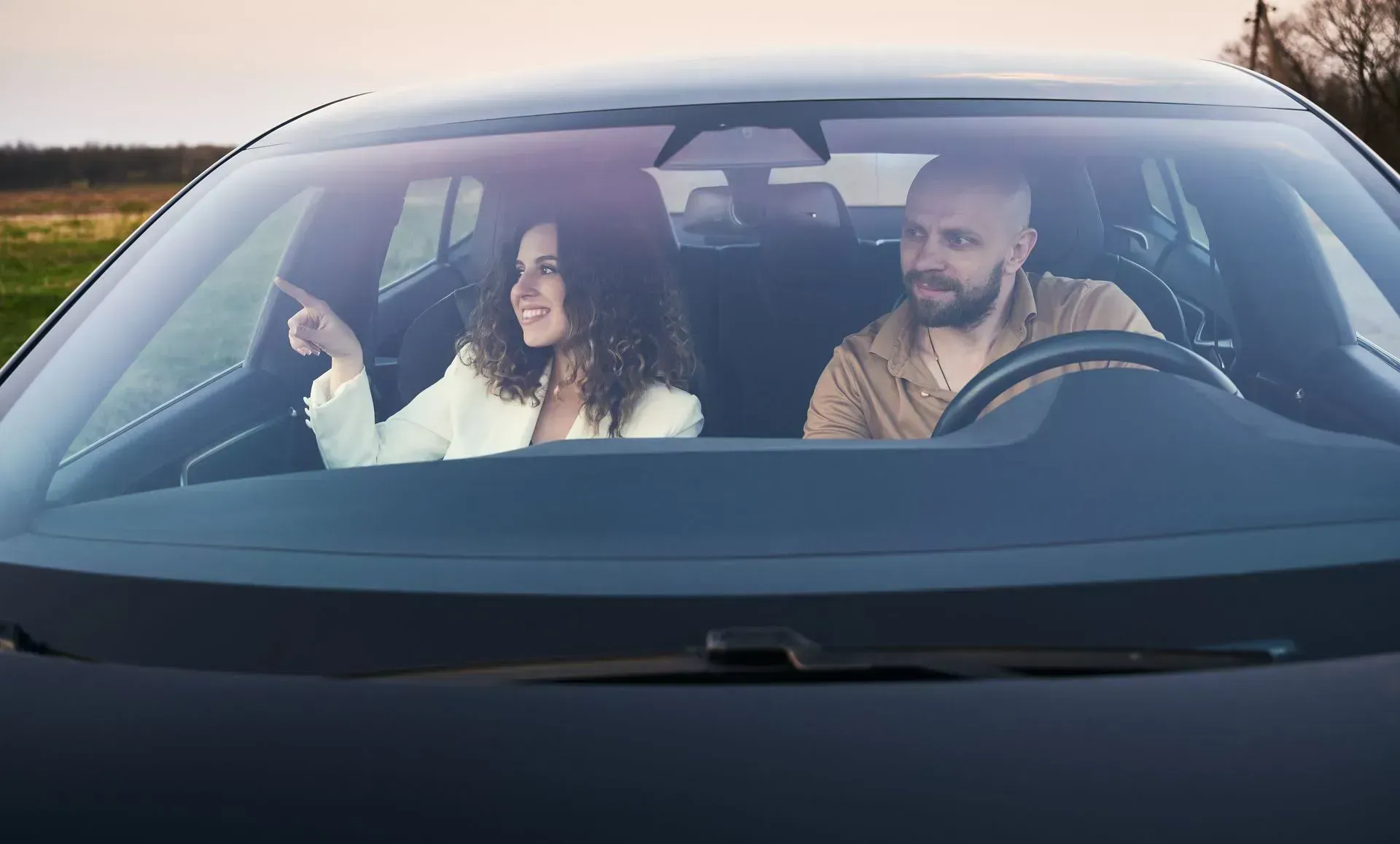A person in a white blazer points through the windshield of a car while sitting next to the driver in a rural setting.