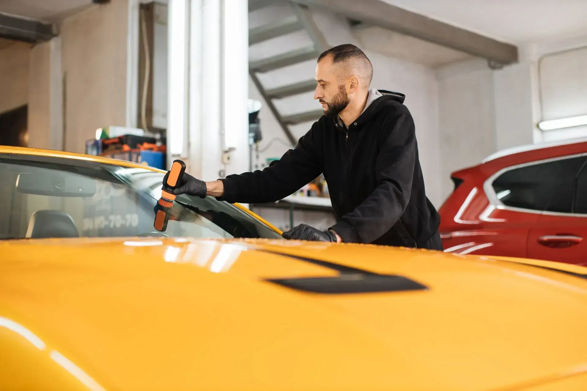 A technician in a black hoodie uses a power tool to polish the windshield of a yellow car in an auto shop.
