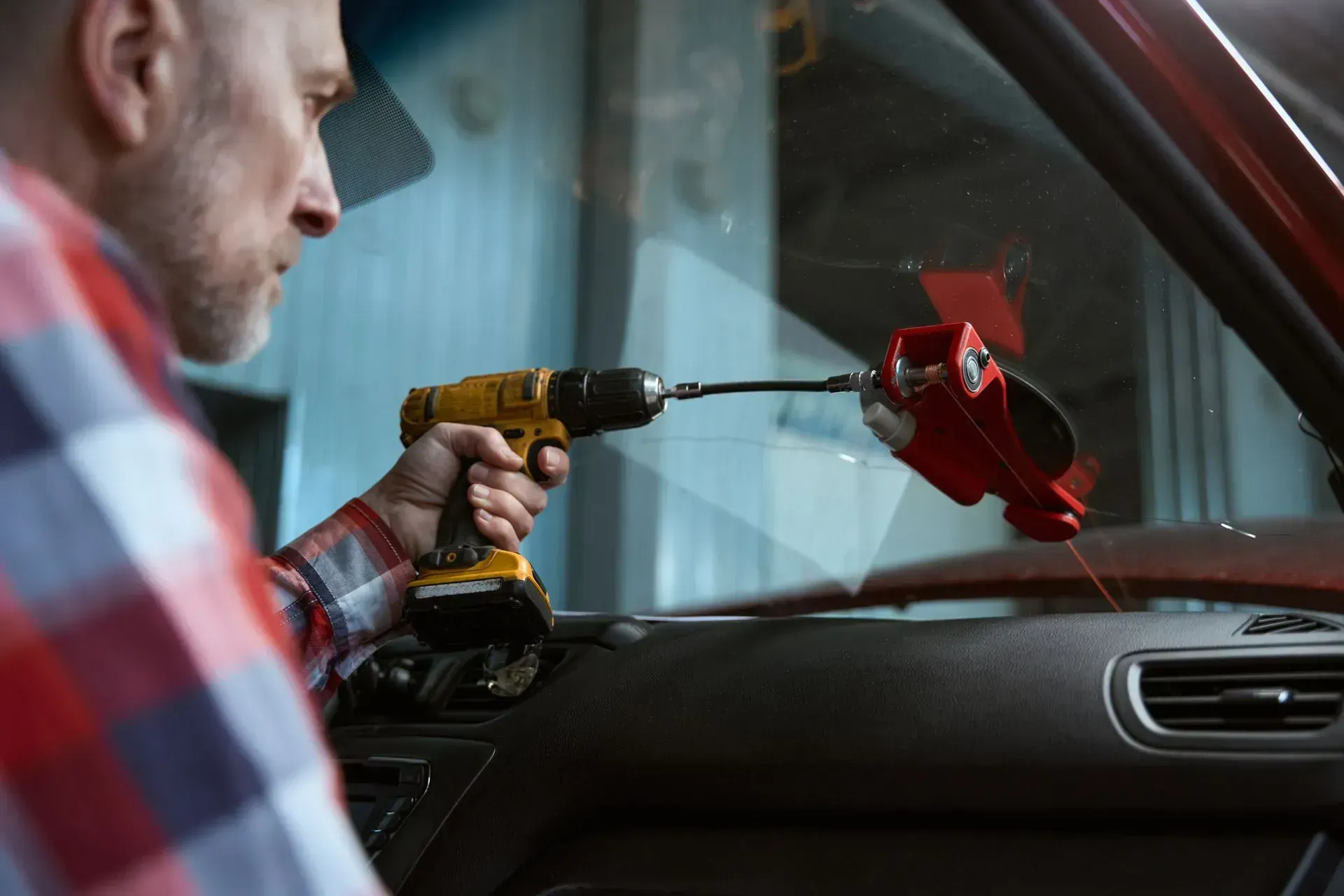 A person in a plaid shirt uses a power drill to operate a red glass repair tool on a vehicle's windshield.