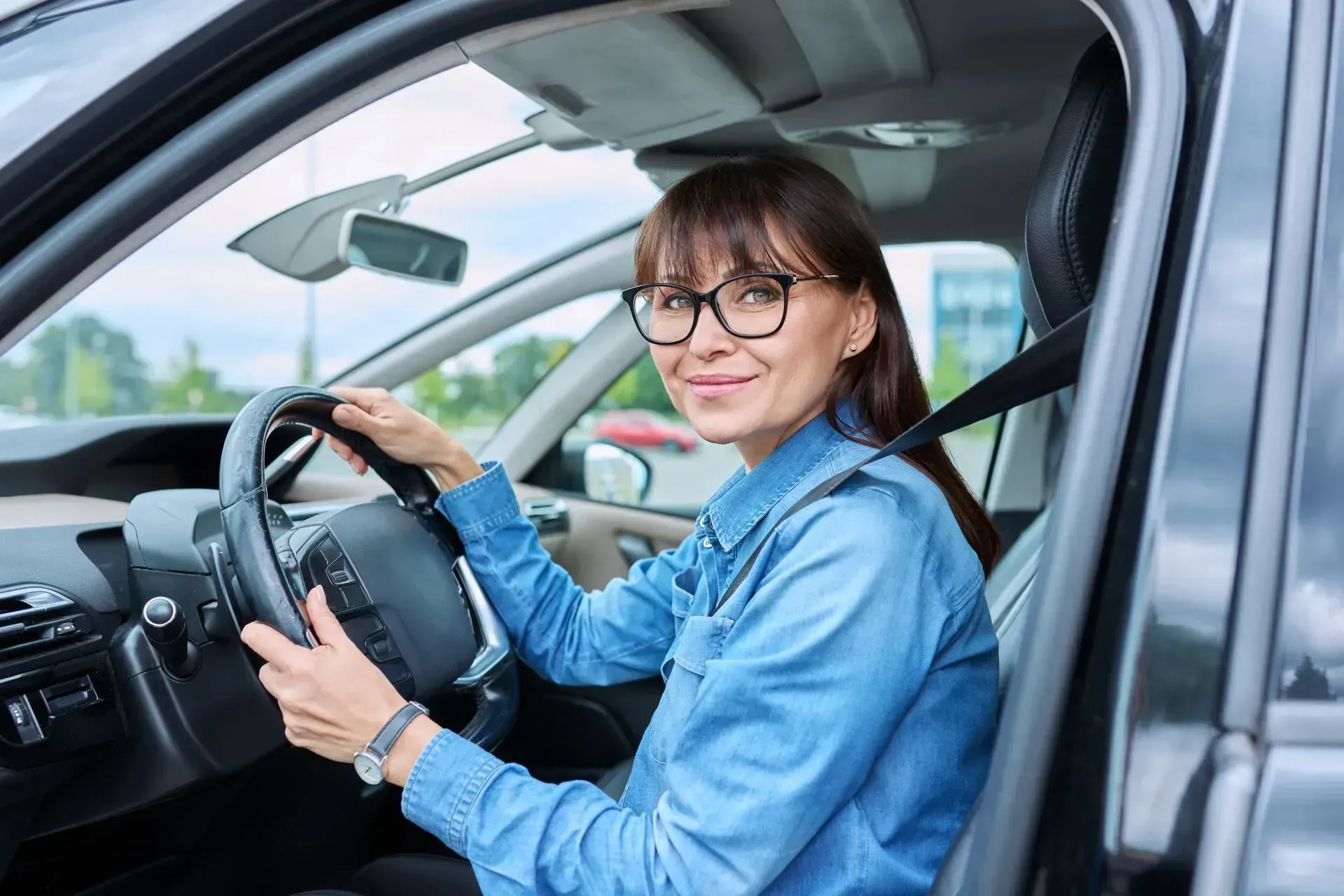 A person with glasses and a denim shirt sitting in the driver's seat of a car, smiling and holding the steering wheel.