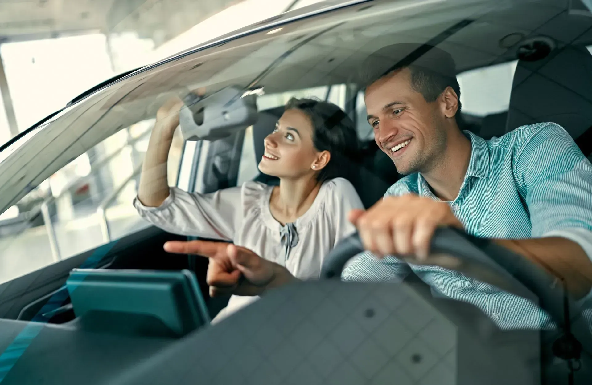 A person adjusts a car's rearview mirror while another person points at the dashboard inside a brightly lit car dealership.
