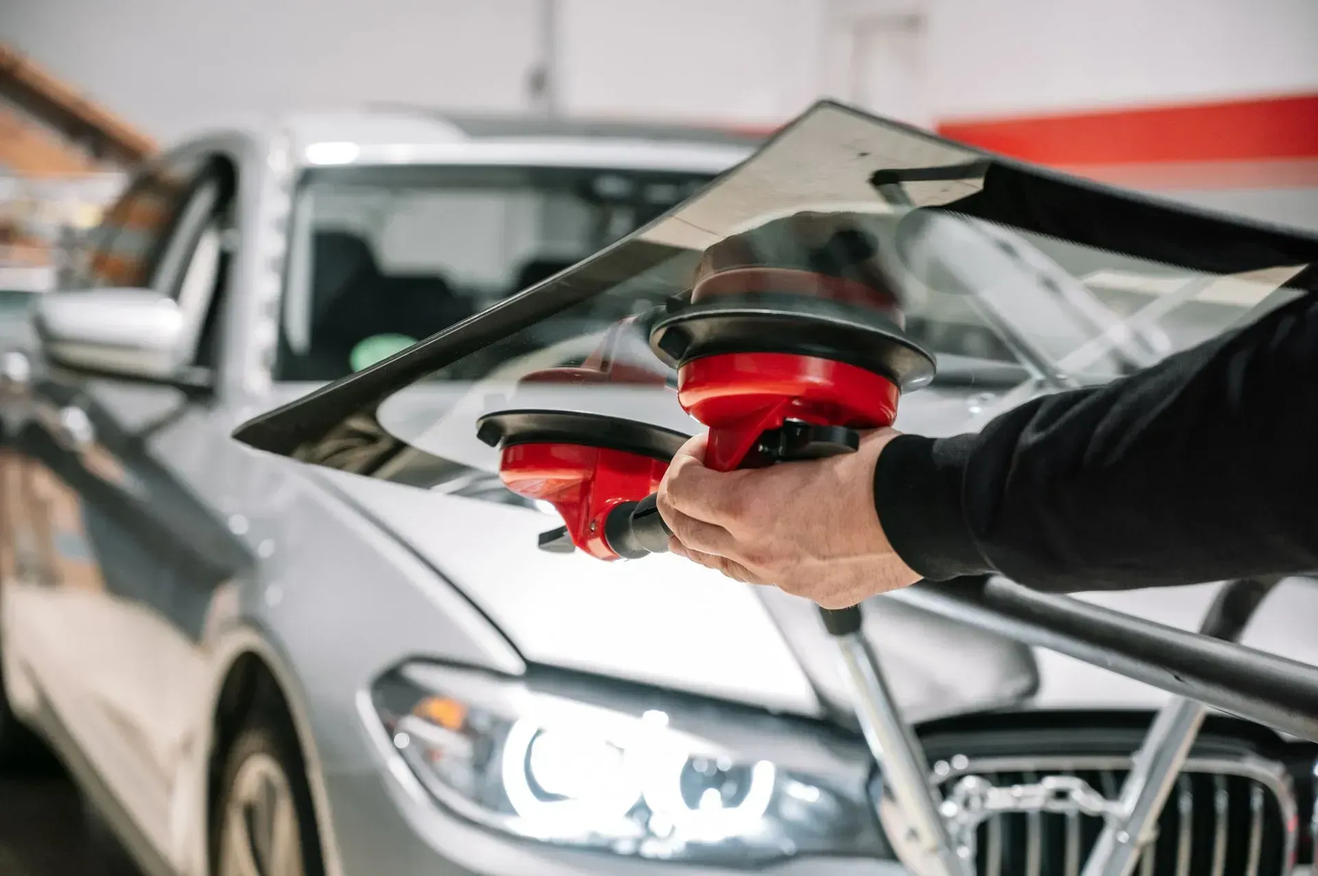 A person holds a car windshield with a red suction cup tool in an automotive garage.