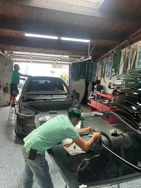 Two auto technicians prepare a replacement windshield inside a garage where another vehicle is being serviced.