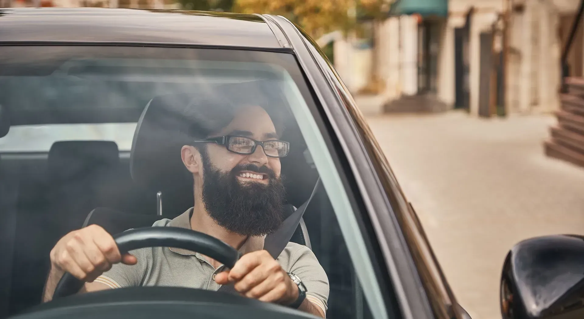 Man with glasses and beard driving a car, smiling. Sunlight on the windshield.