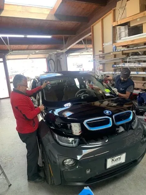 Two technicians work on the windshield of a black BMW i3 inside a bright workshop.