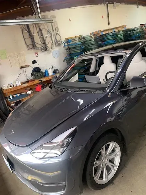 A gray Tesla Model Y with a missing windshield parked inside an automotive workshop with workbenches and equipment.