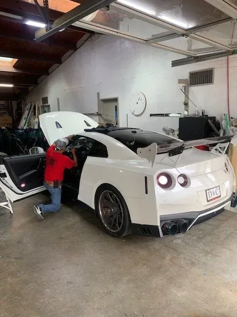 A person in a red shirt kneels inside a white Nissan GT-R sports car with an open hood inside a workshop.