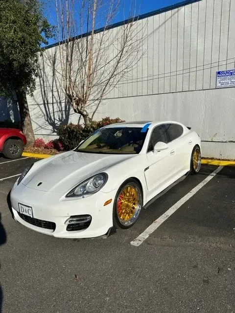 A white Porsche Panamera with gold rims parked in a paved lot next to a white building.