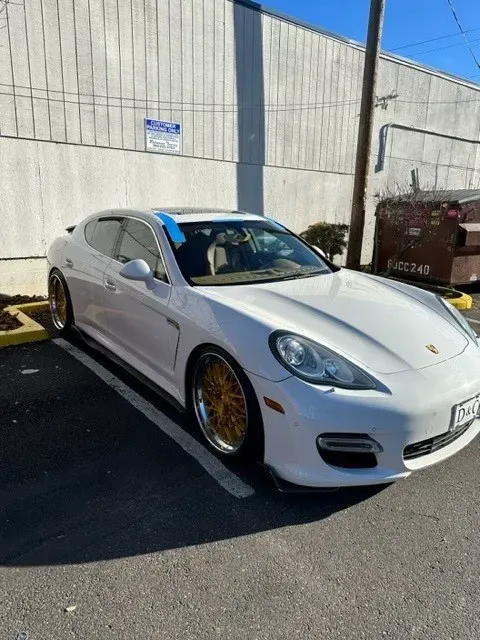 White Porsche Panamera with gold rims parked in a lot next to a grey building and a dumpster on a sunny day.