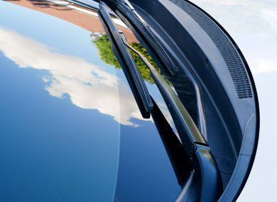 Windshield with wiper blades and reflections of sky and building.