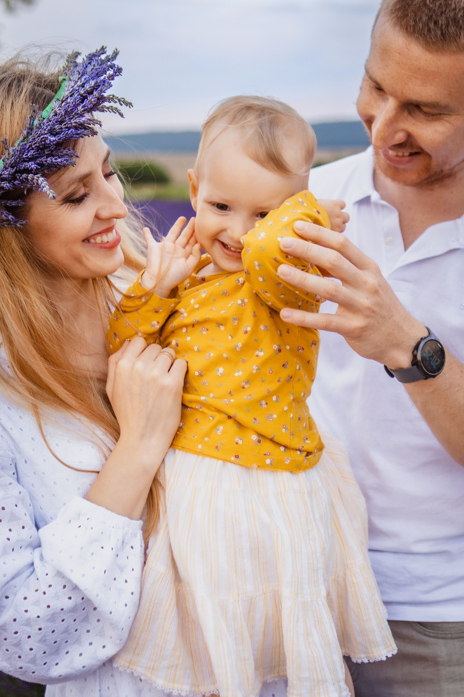Parents holding a smiling toddler in a lavender field; the child is wearing a yellow shirt and cream skirt.