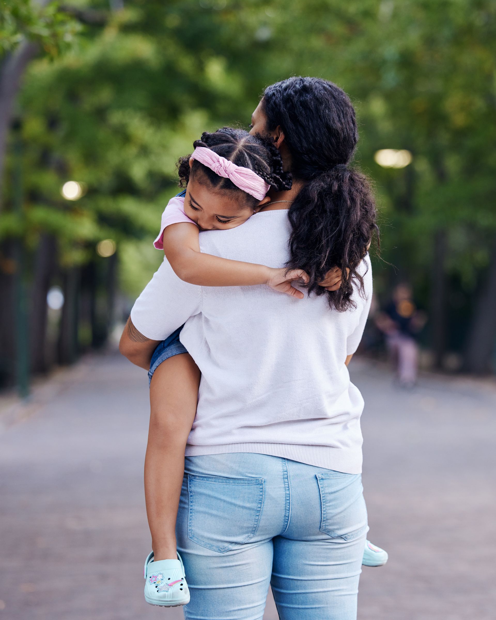 Woman carrying a young girl outdoors on a path lined with trees. The girl hugs the woman.