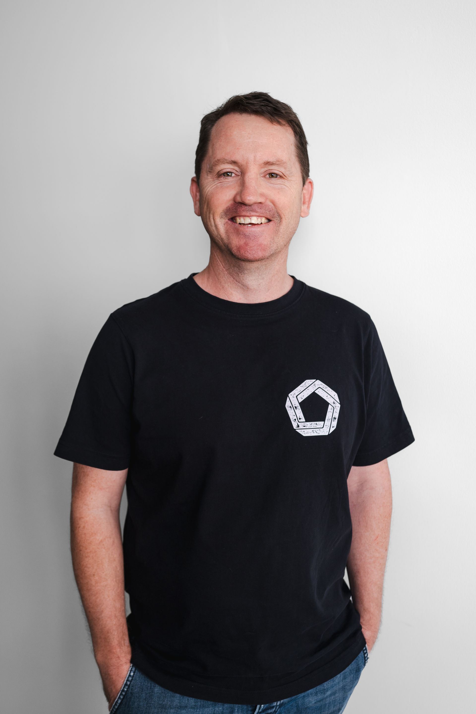 A man wearing a black t-shirt and jeans is standing with his hands in his pockets.