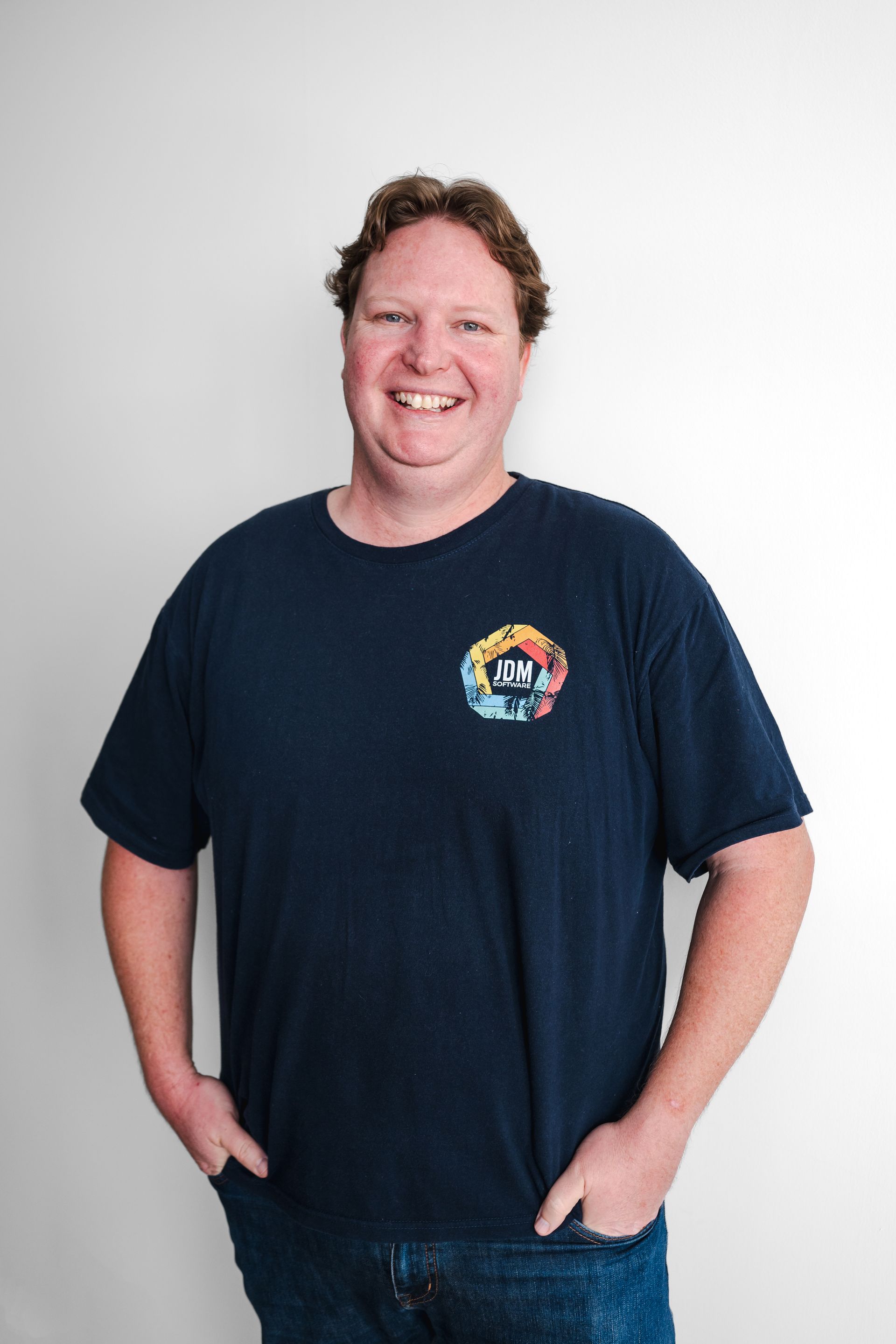 A man wearing a blue t-shirt is smiling and standing with his hands in his pockets.