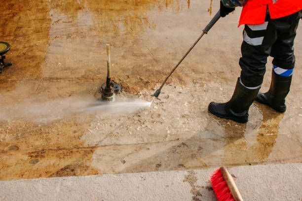 A man in an orange vest and safety boots cleans a concrete floor during post-flood cleaning and restoration efforts.
