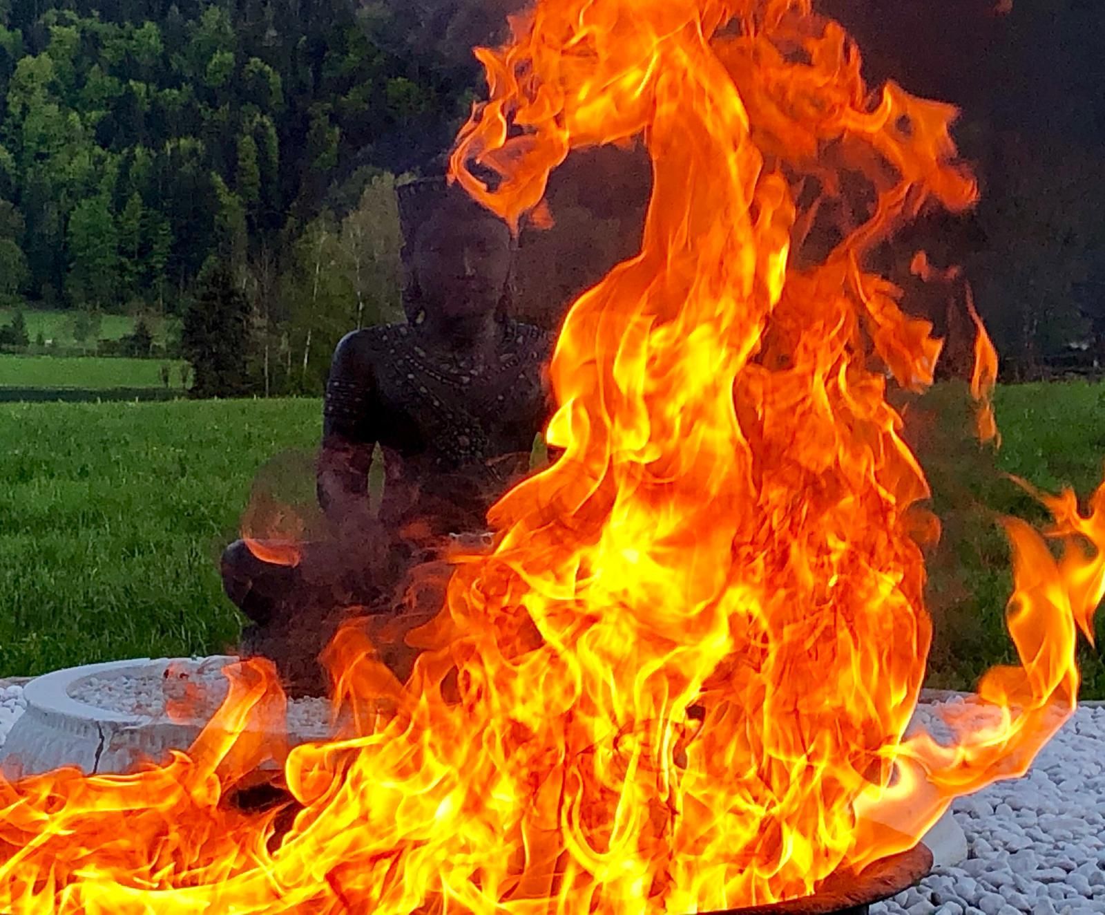 Flammen umhüllen eine dunkle Statue in einer Feuerstelle, die auf einem grasbewachsenen Feld mit Bäumen im Hintergrund steht.