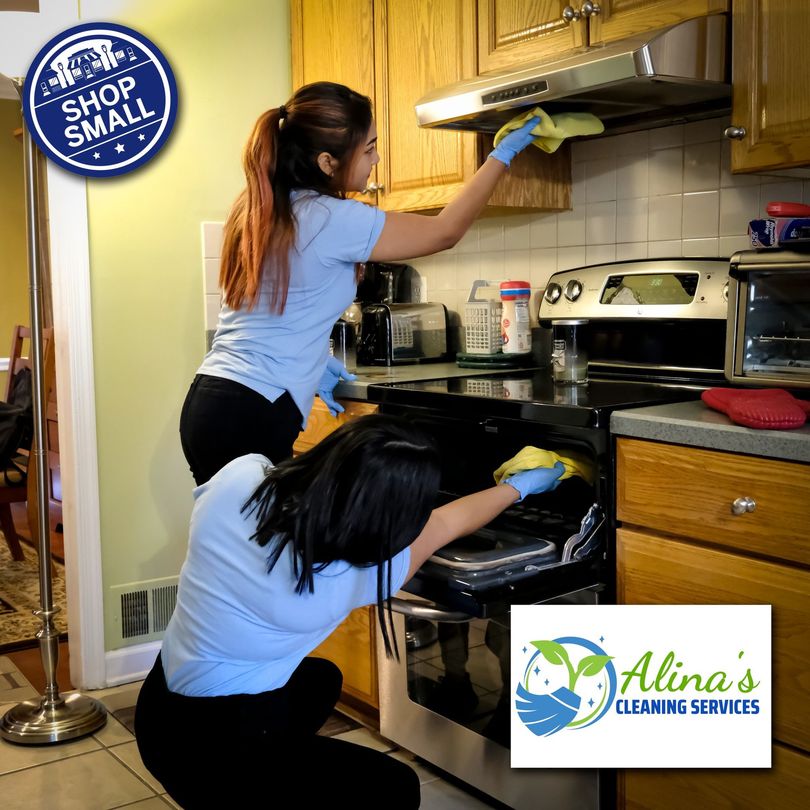 Two women cleaning a kitchen; one wiping the range hood, the other cleaning the oven.