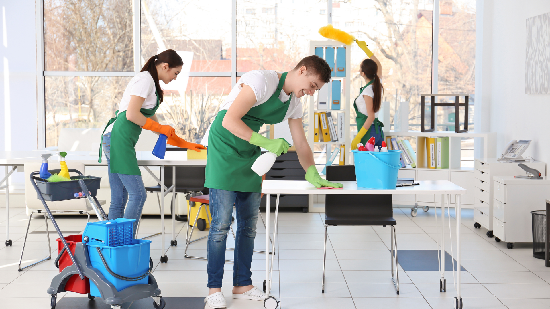 Three office cleaners wiping desks, wearing green aprons and gloves, in a bright office.