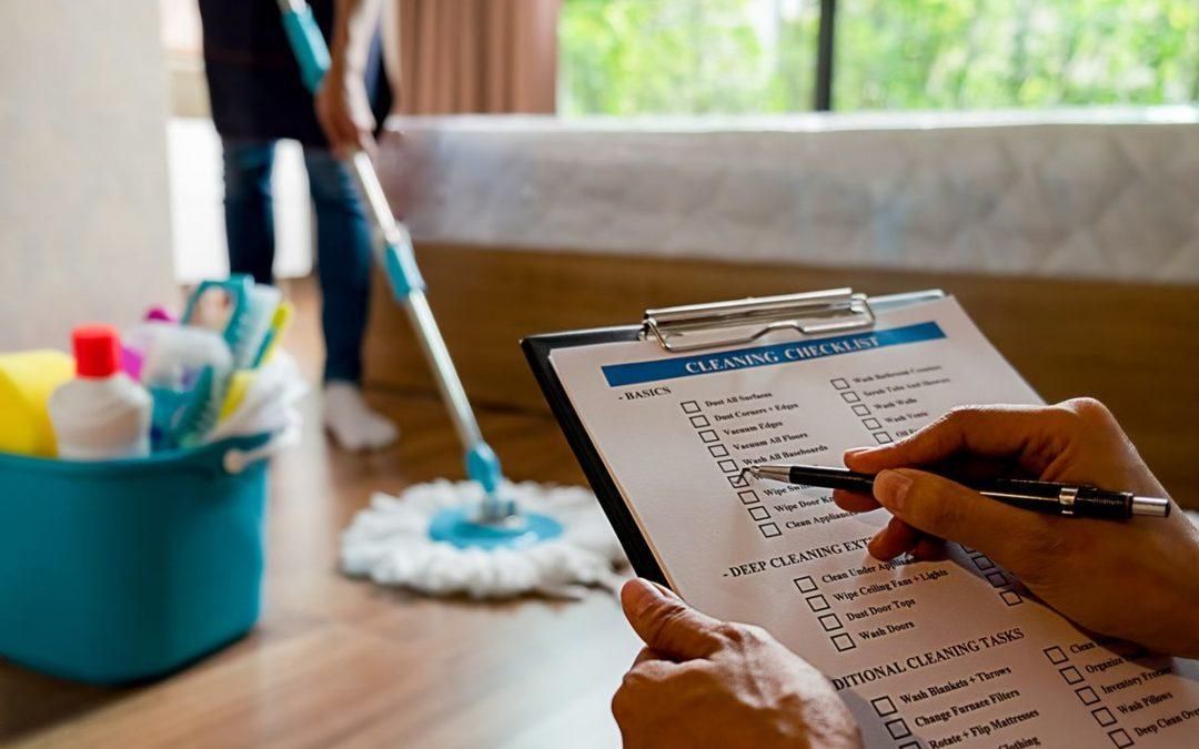 A person checks a cleaning checklist on a clipboard while someone mops the floor nearby, with a bucket of supplies.