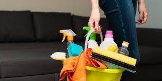 Person holding a yellow bucket filled with cleaning supplies near a dark gray couch.