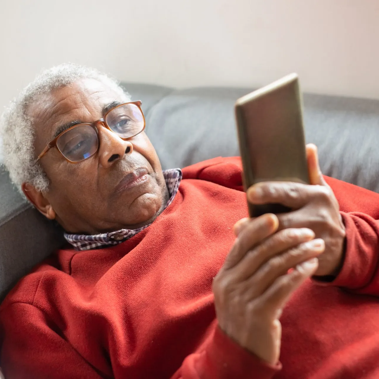 An elderly man in a red jersey, lying on the sofa and looking at a smart phone