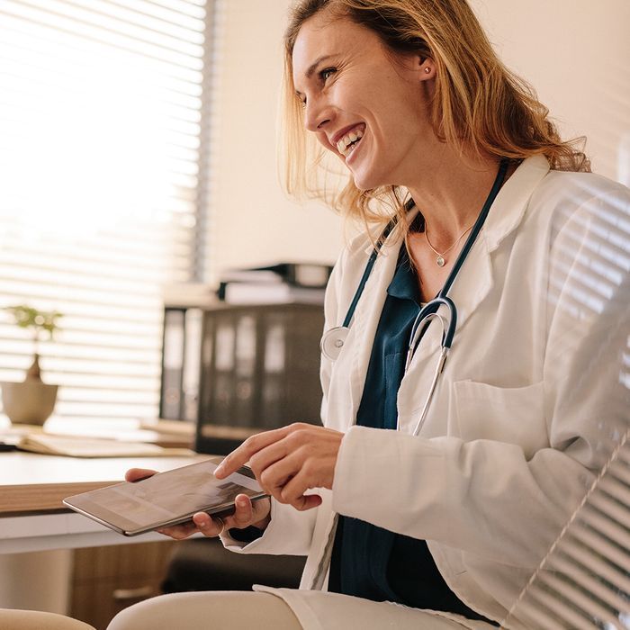 A female doctor is sitting at a desk holding a tablet and smiling.