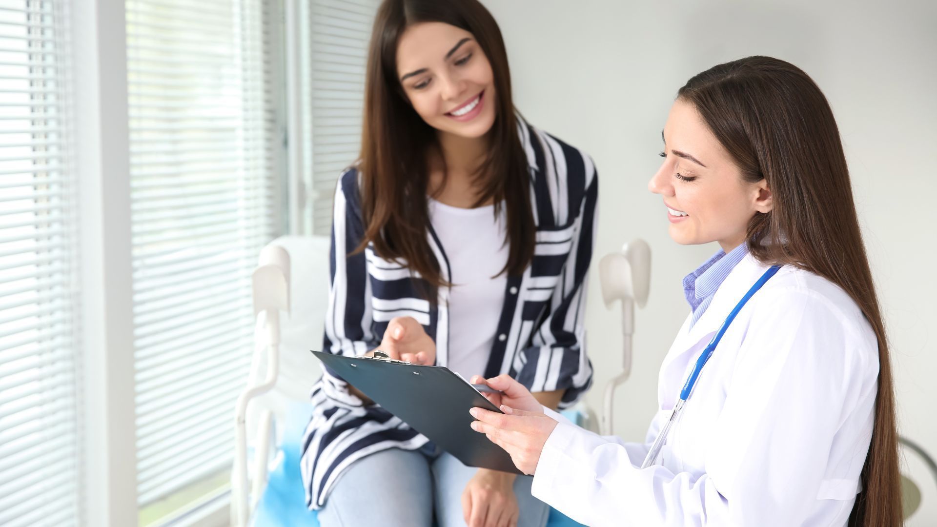 A woman is sitting in a chair talking to a doctor who is holding a clipboard.