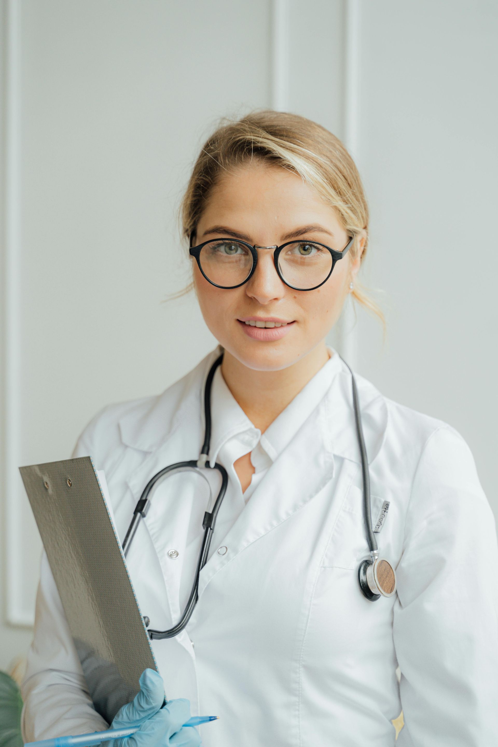 A female doctor wearing glasses and a stethoscope is holding a clipboard.