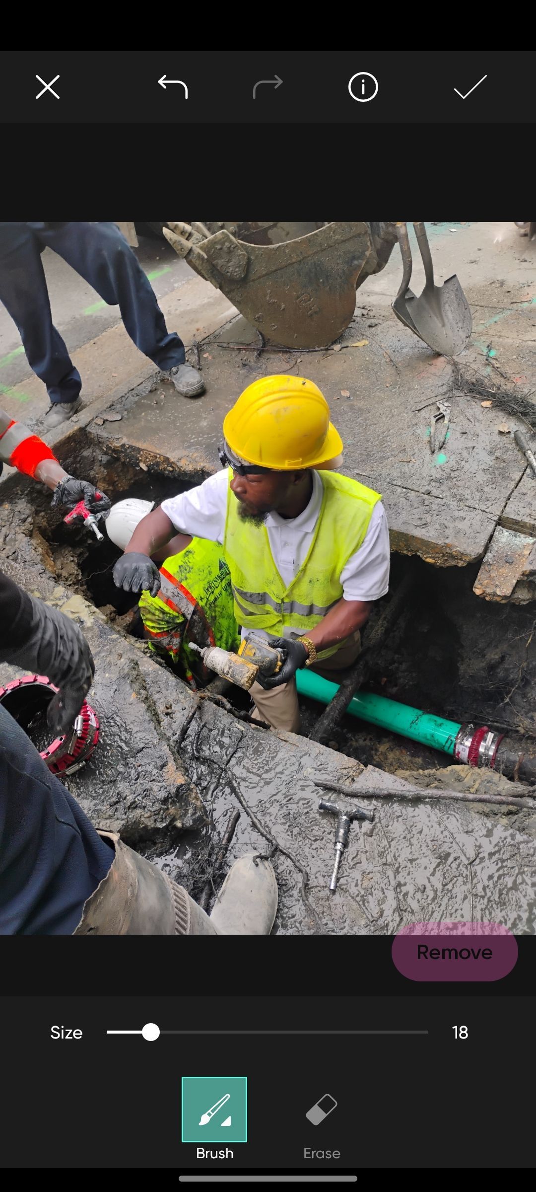 A worker wearing a yellow hard hat and safety vest repairs a green pipe in an excavated trench.