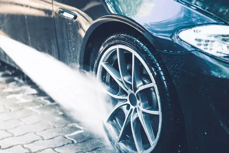 A jet of water from a pressure washer cleans the wheel and tire of a dark-colored car parked on a paved driveway.
