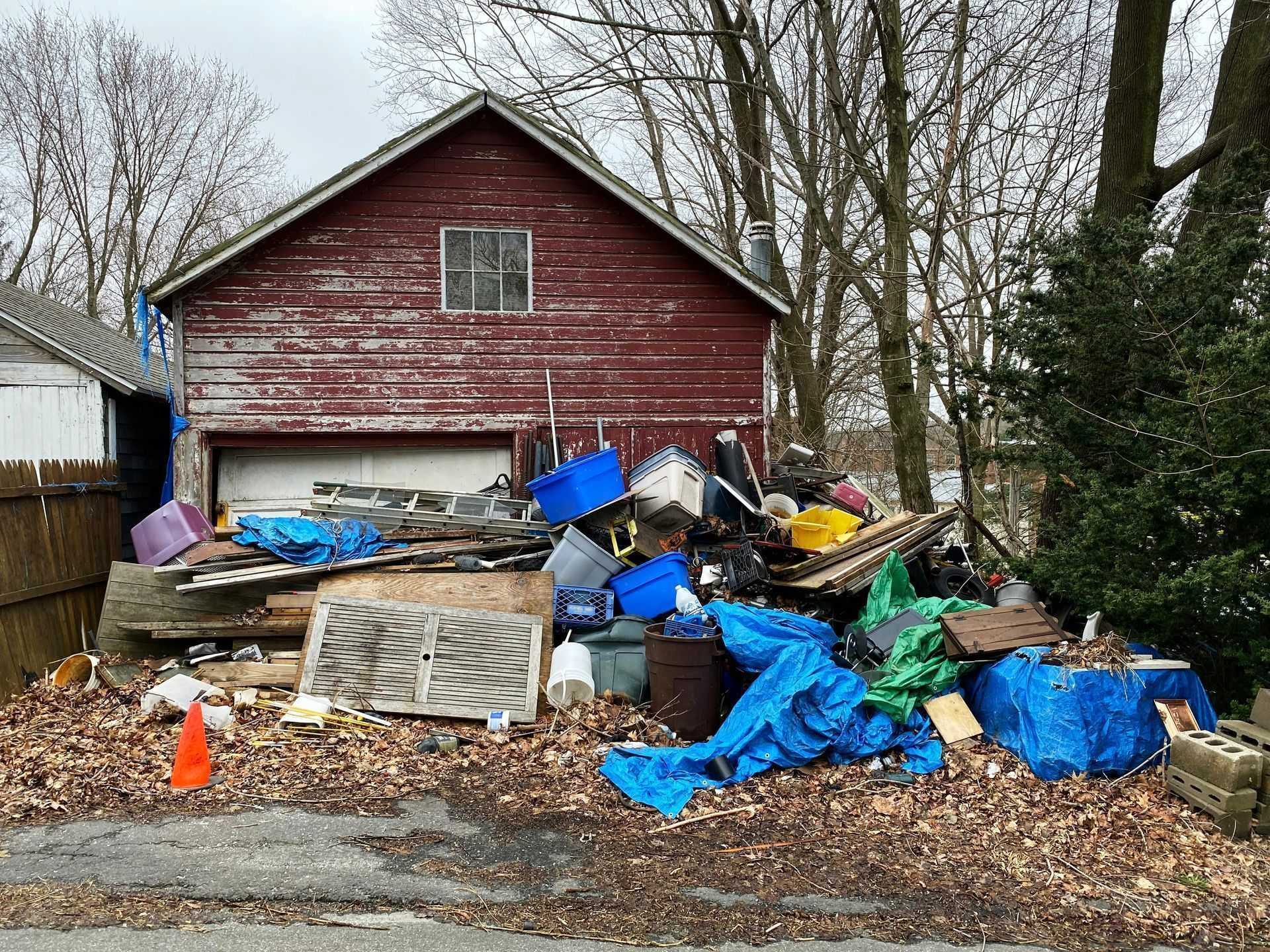 A red garage with a large pile of miscellaneous debris, trash, and blue tarps scattered in the yard in front of it.