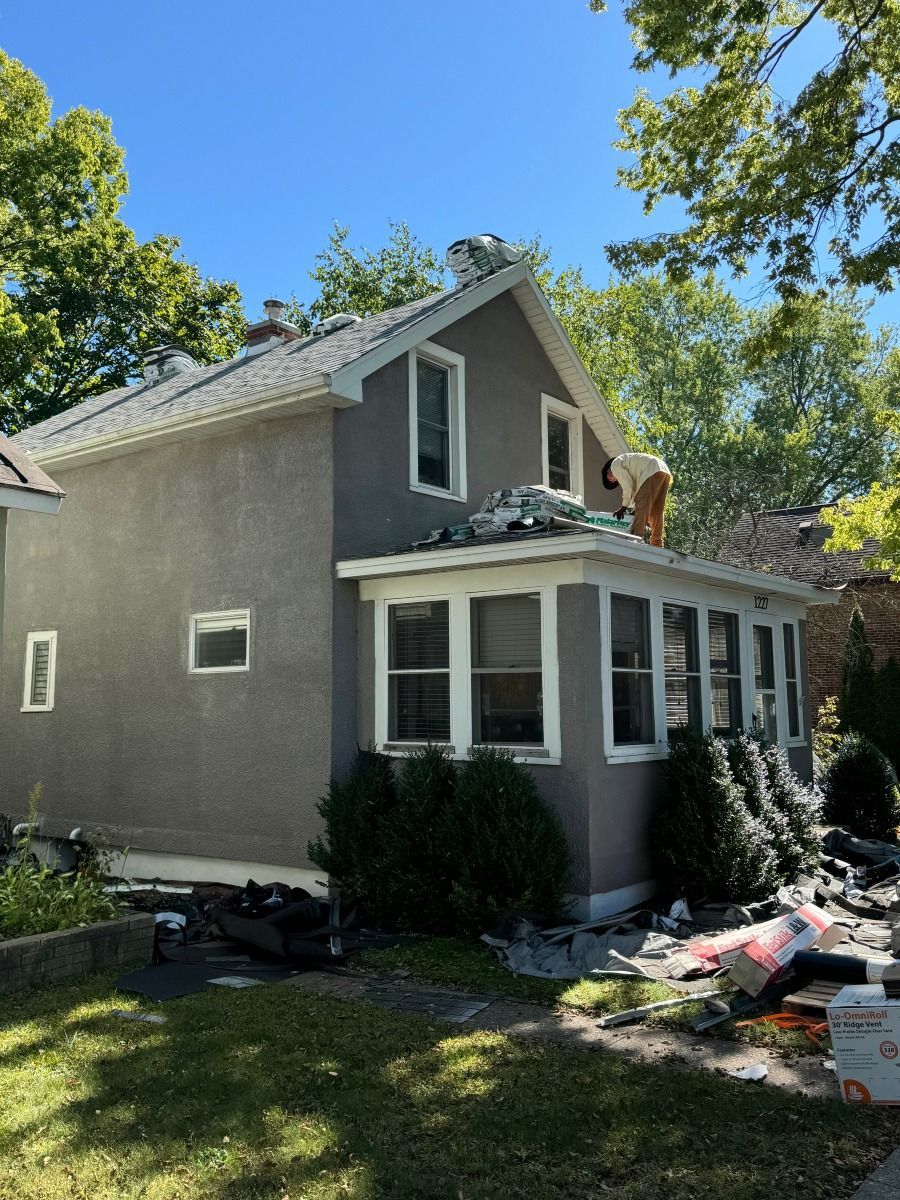 Workers replace roof shingles on a two-story gray house under a bright blue sky.