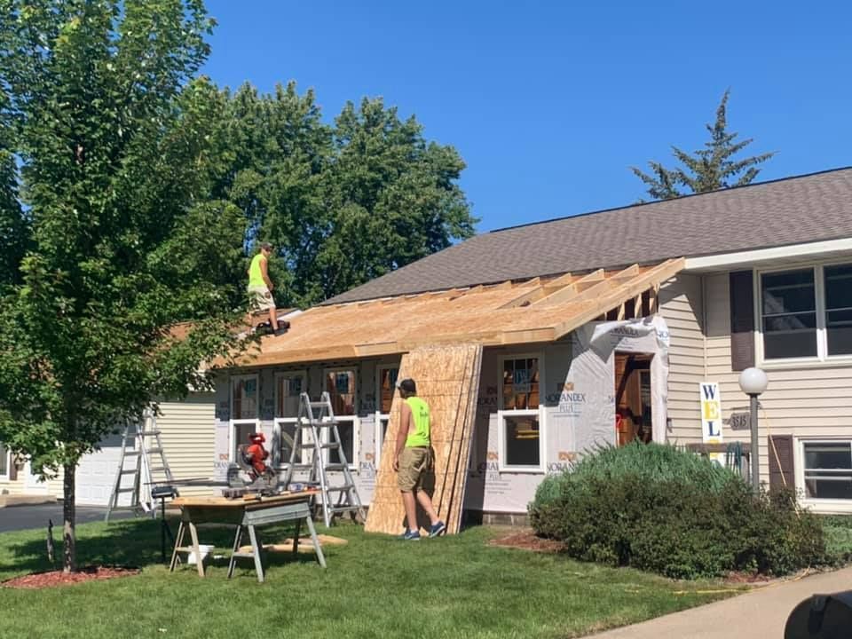 Workers on a roof removing shingles, sunny day. House with open roof. Tools and materials on the ground.