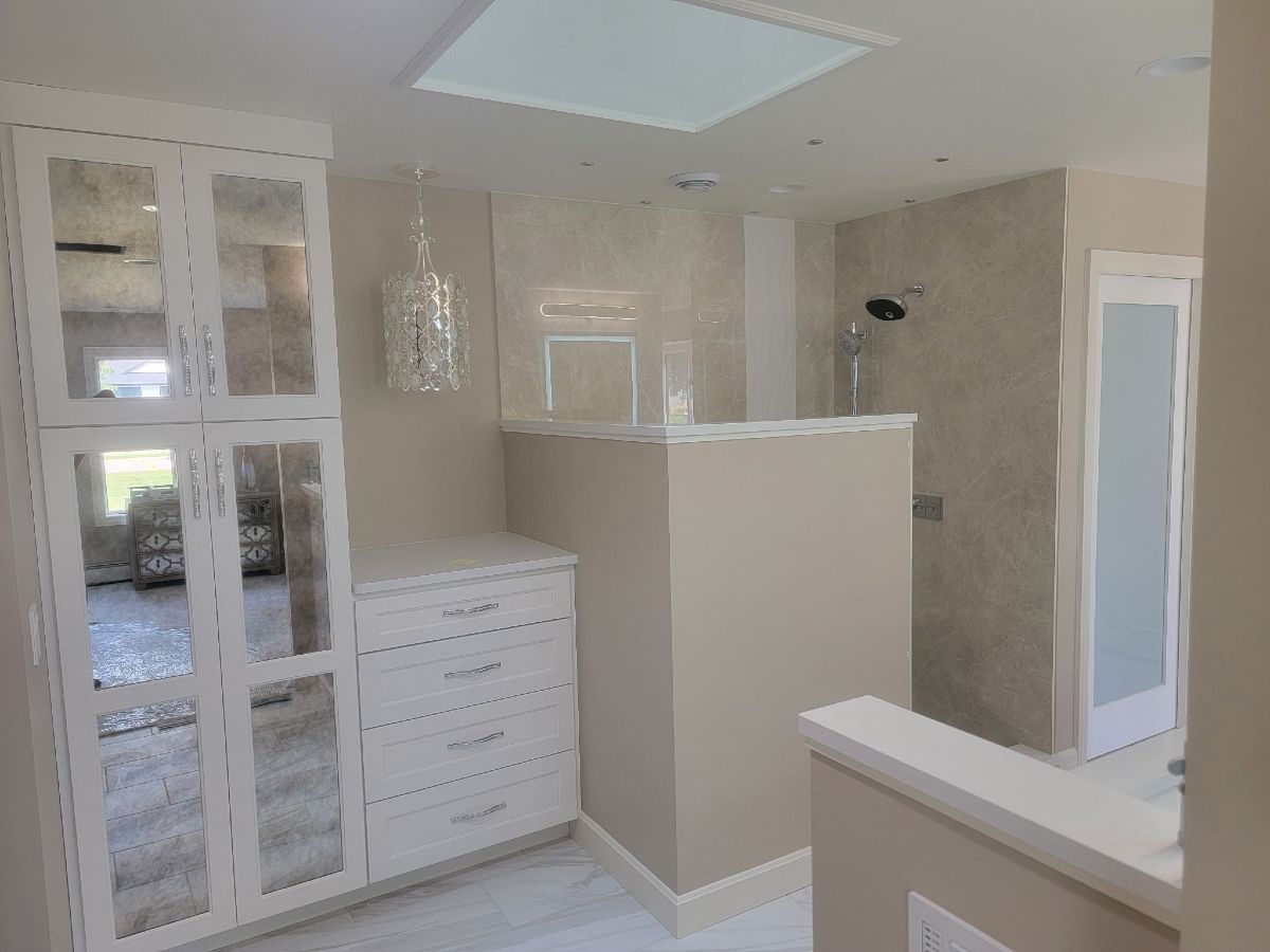 A bright, modern bathroom featuring a white wardrobe with mirrored panels, a dresser, and a walk-in shower with stone tile.