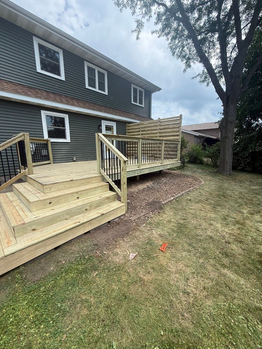 A new wooden deck with stairs and privacy fencing attached to the side of a grey two-story house.