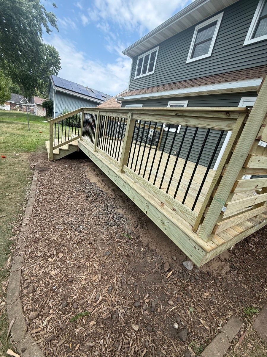A newly built wooden deck with black metal balusters attached to the side of a grey house, featuring a small staircase.