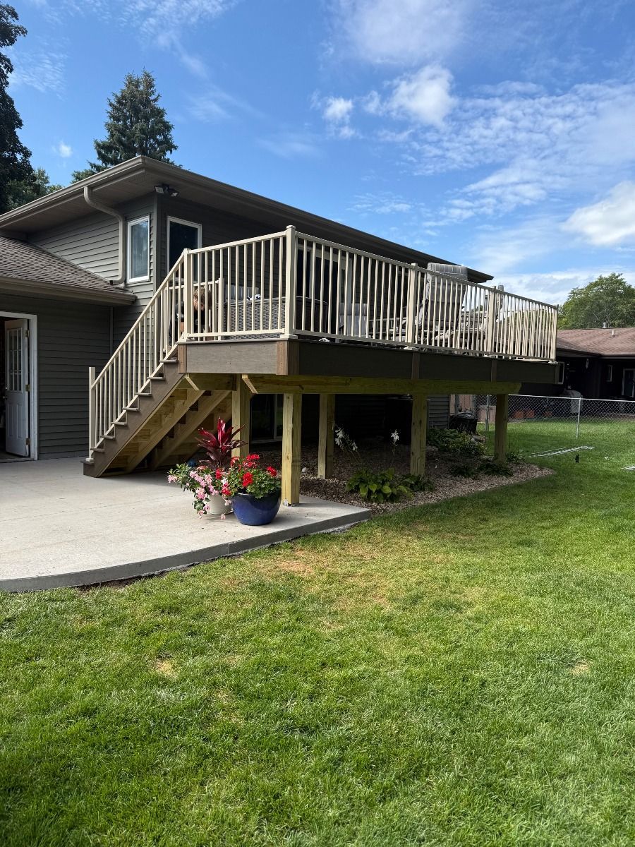 A tan-colored wooden deck with white railings attached to the side of a grey house, above a concrete patio and green lawn.