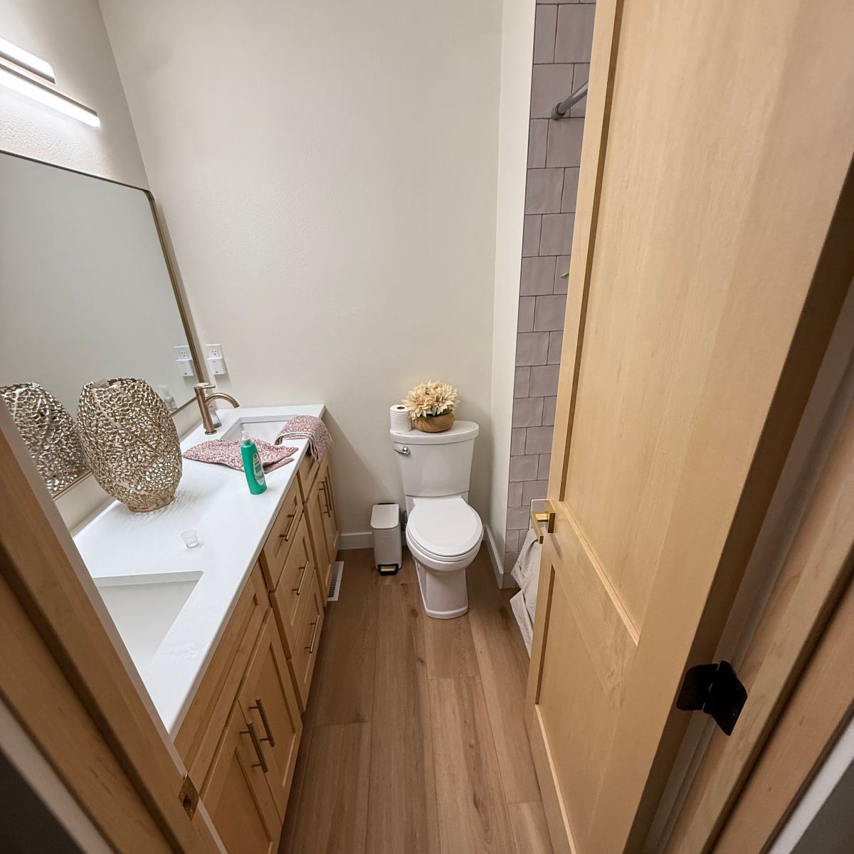 A bathroom with a wooden vanity, white countertop, mirror, toilet, and light wood-tone flooring.