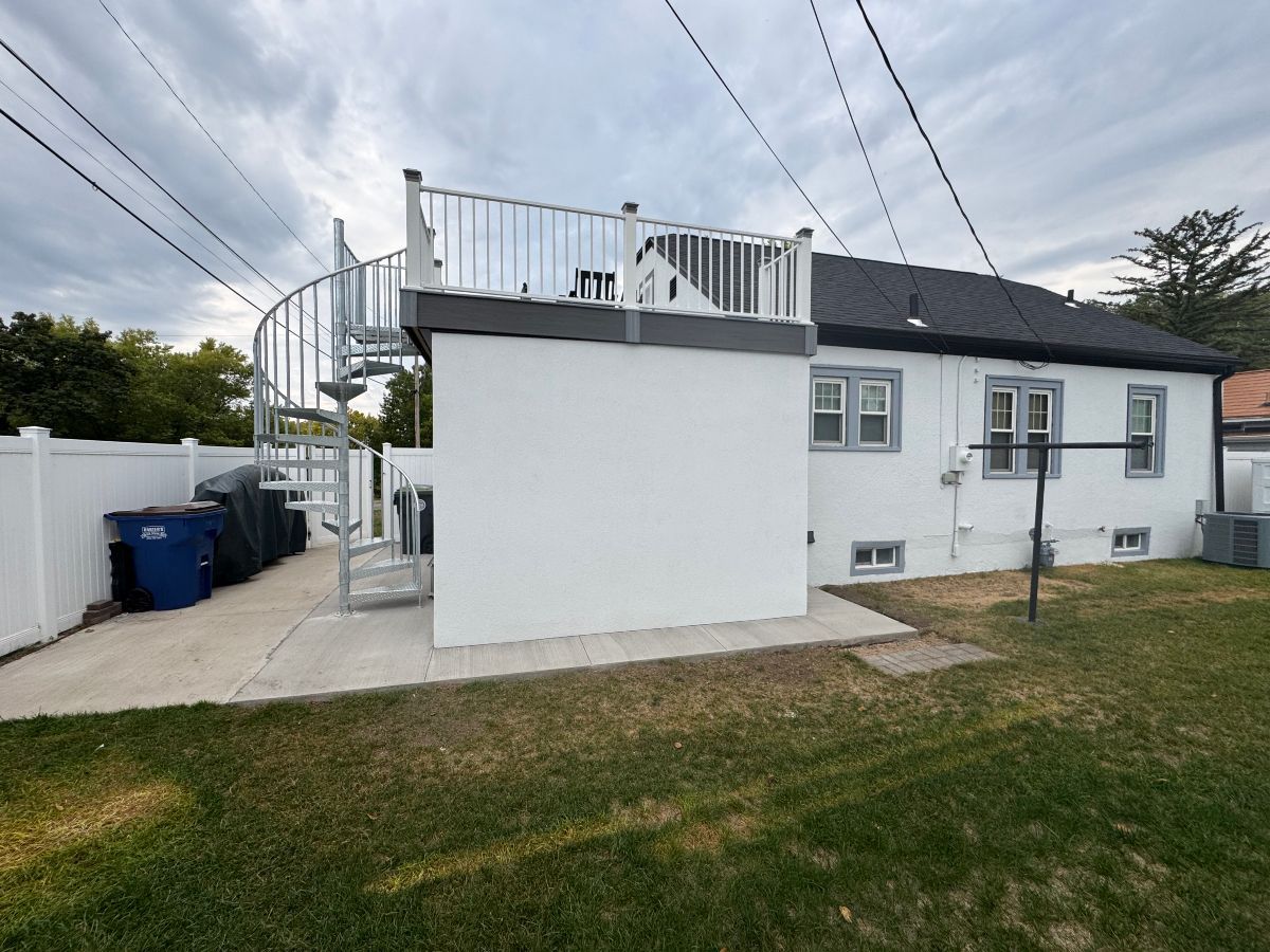 A white house with a rooftop deck accessed by a metal spiral staircase, located in a fenced yard.