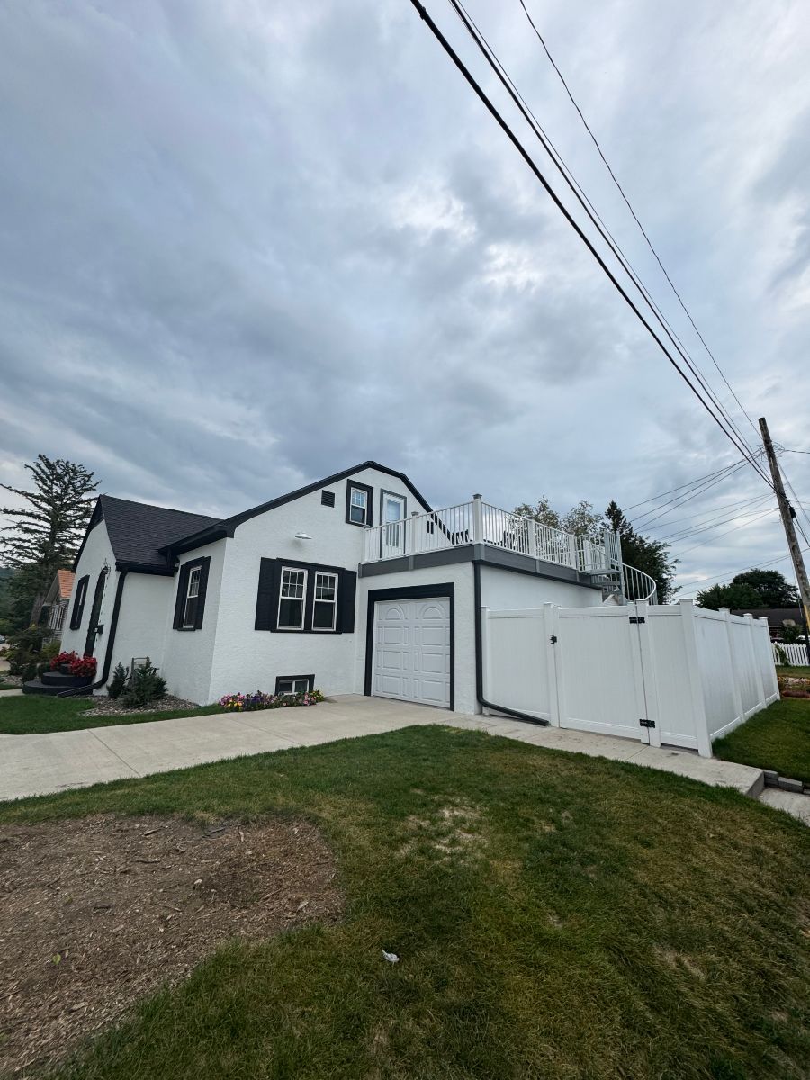 A white house with a dark roof, black trim, a garage, and a fenced deck under a cloudy sky.