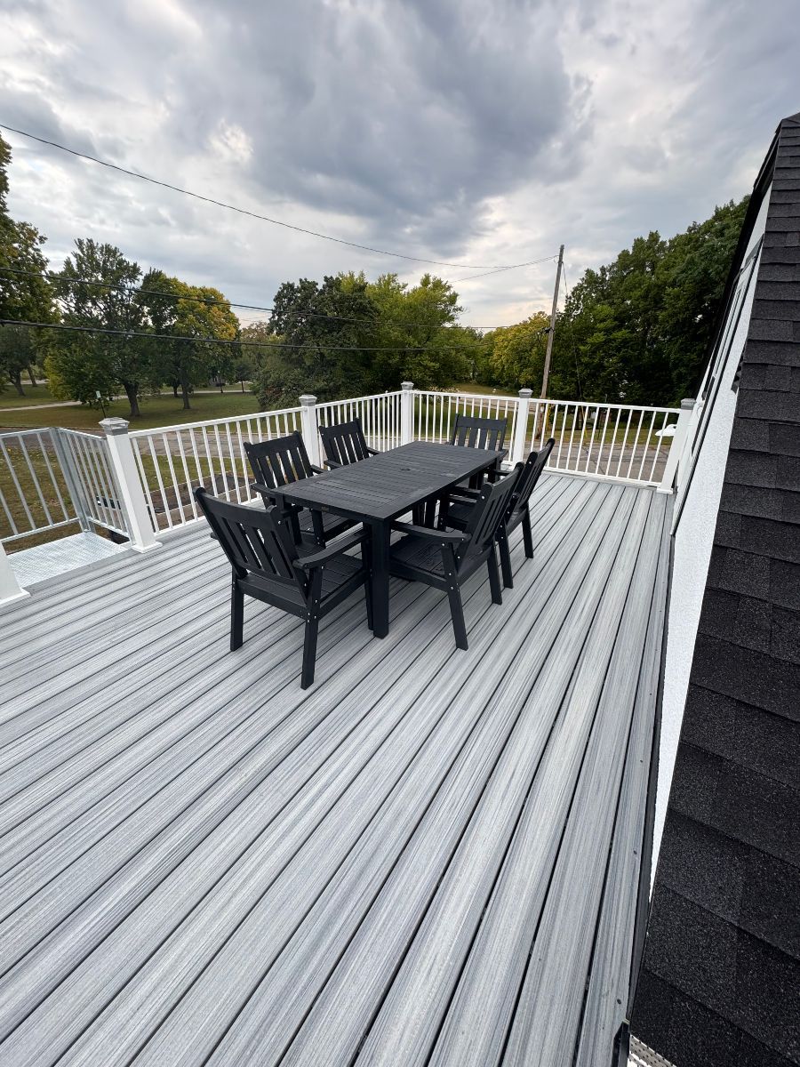 A dining table and chairs on a grey, elevated deck with white railings, set against a backdrop of trees and a cloudy sky.