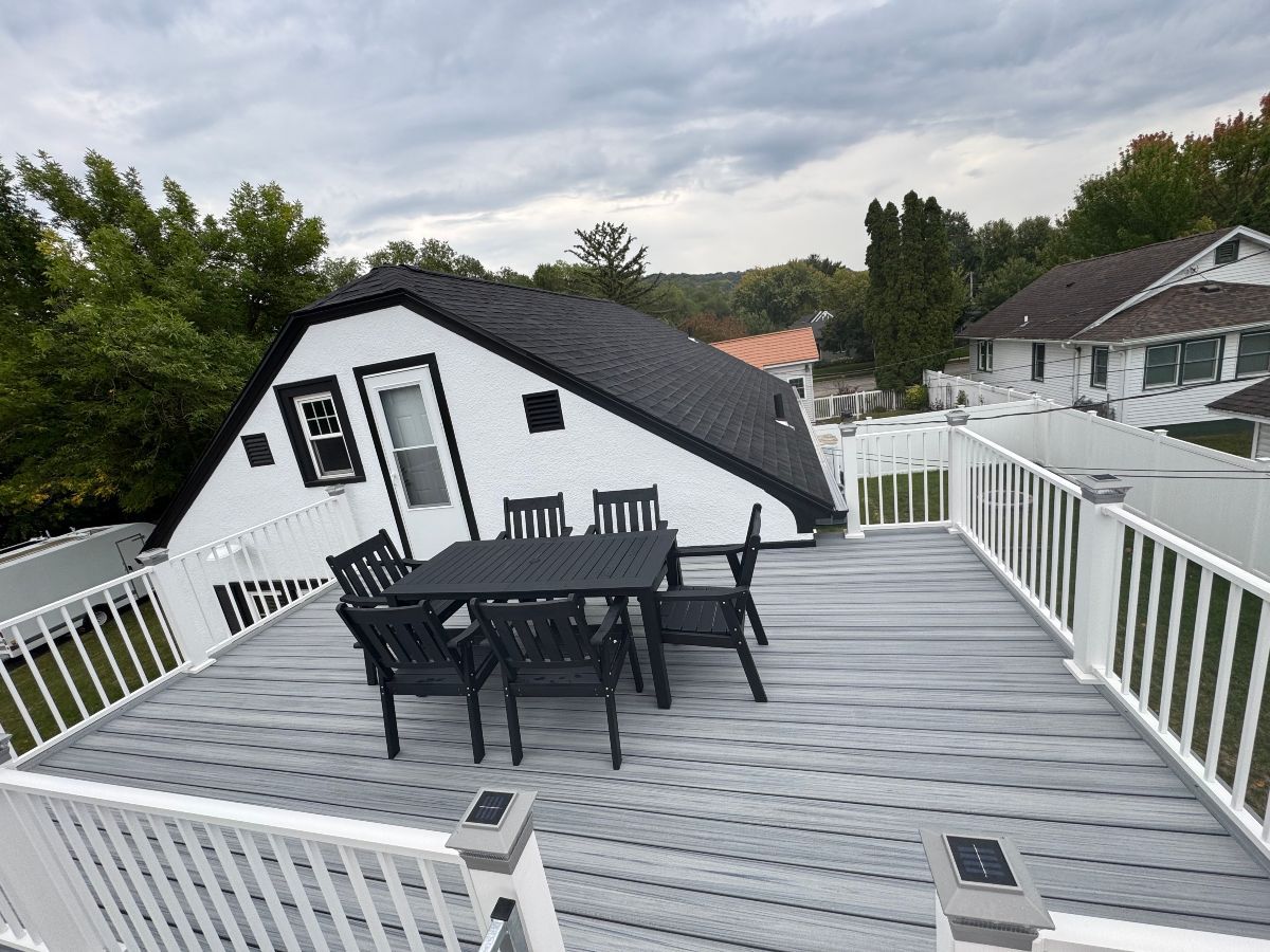 A gray composite deck with a black dining set, surrounded by white railings, attached to a white house with a dark roof.