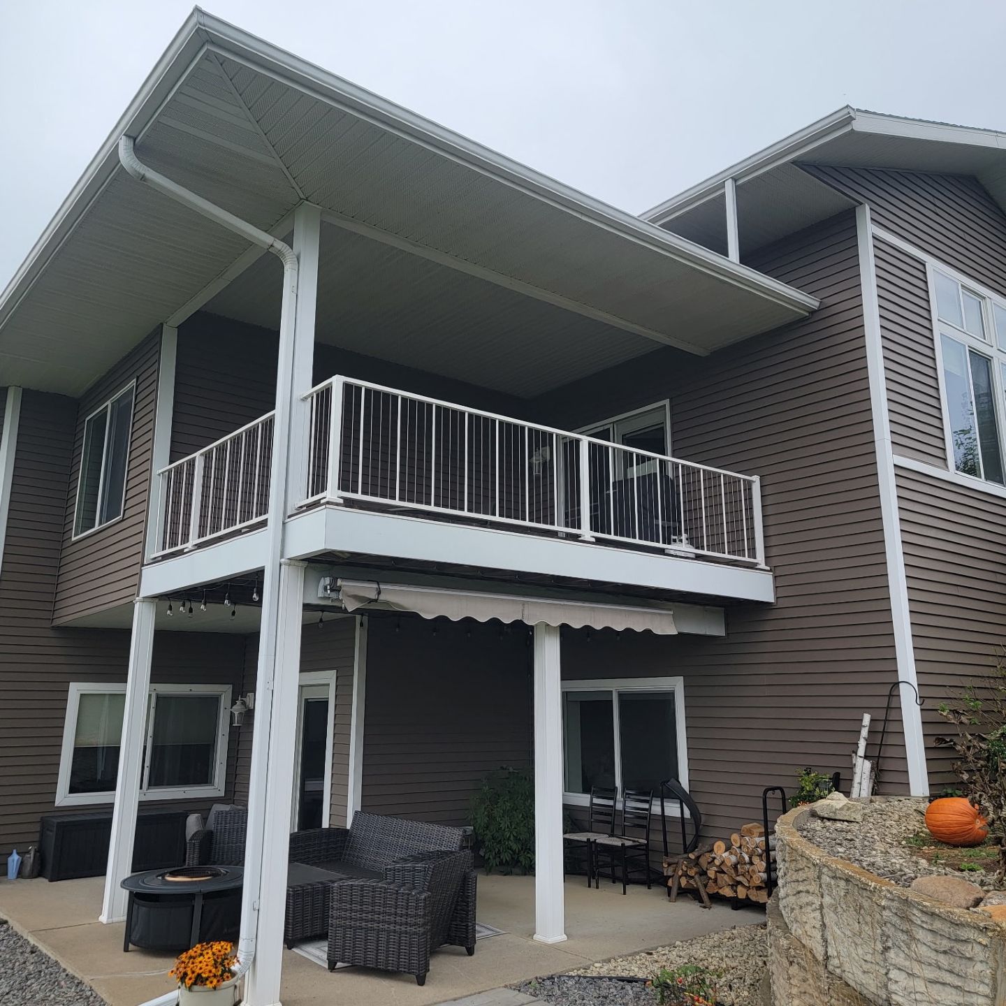 A two-story brown house with a large upper deck, white railings, and a covered lower patio with outdoor furniture.