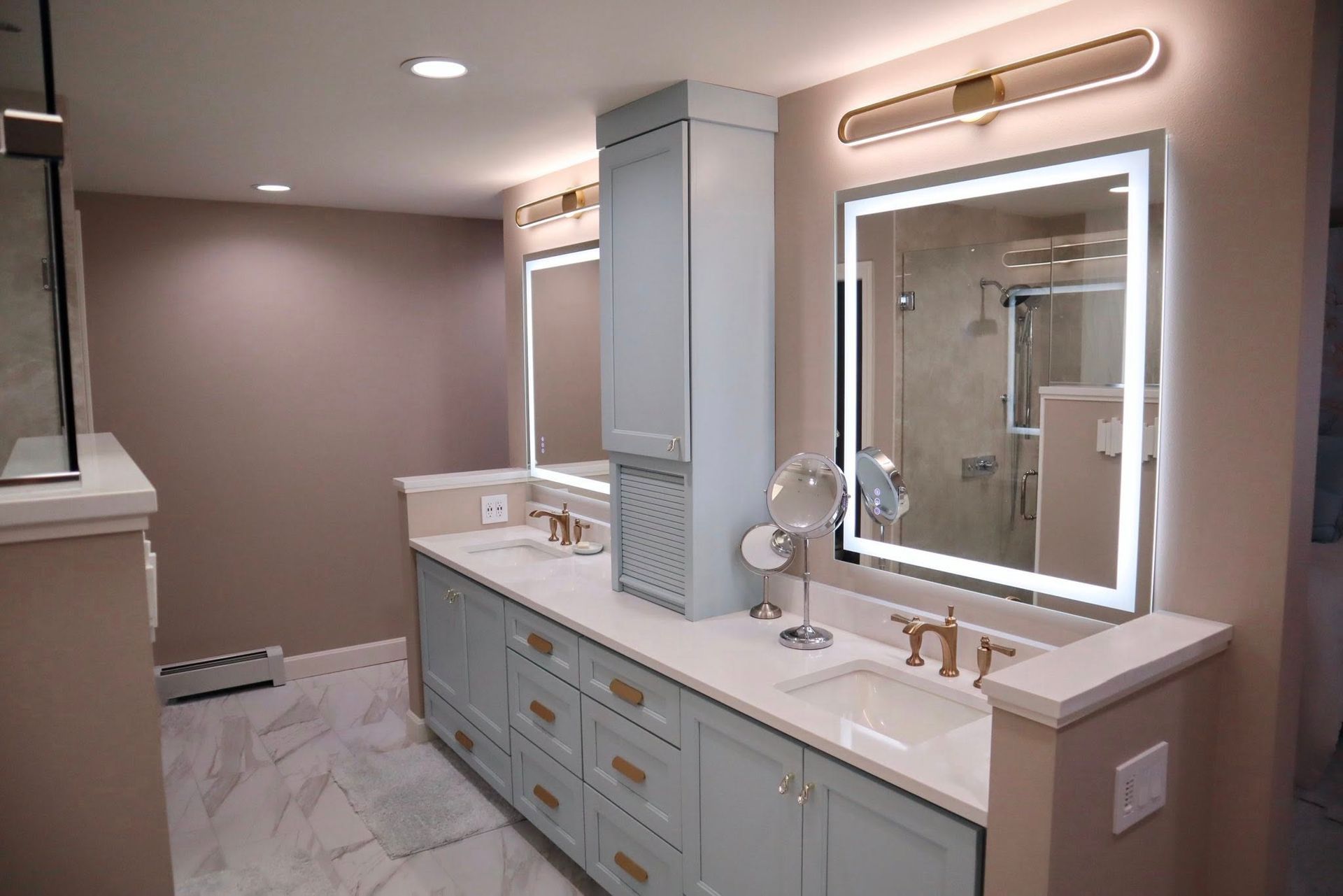 A modern bathroom with a dual vanity, light blue cabinetry, light-up mirrors, gold fixtures, and marbled tile flooring.
