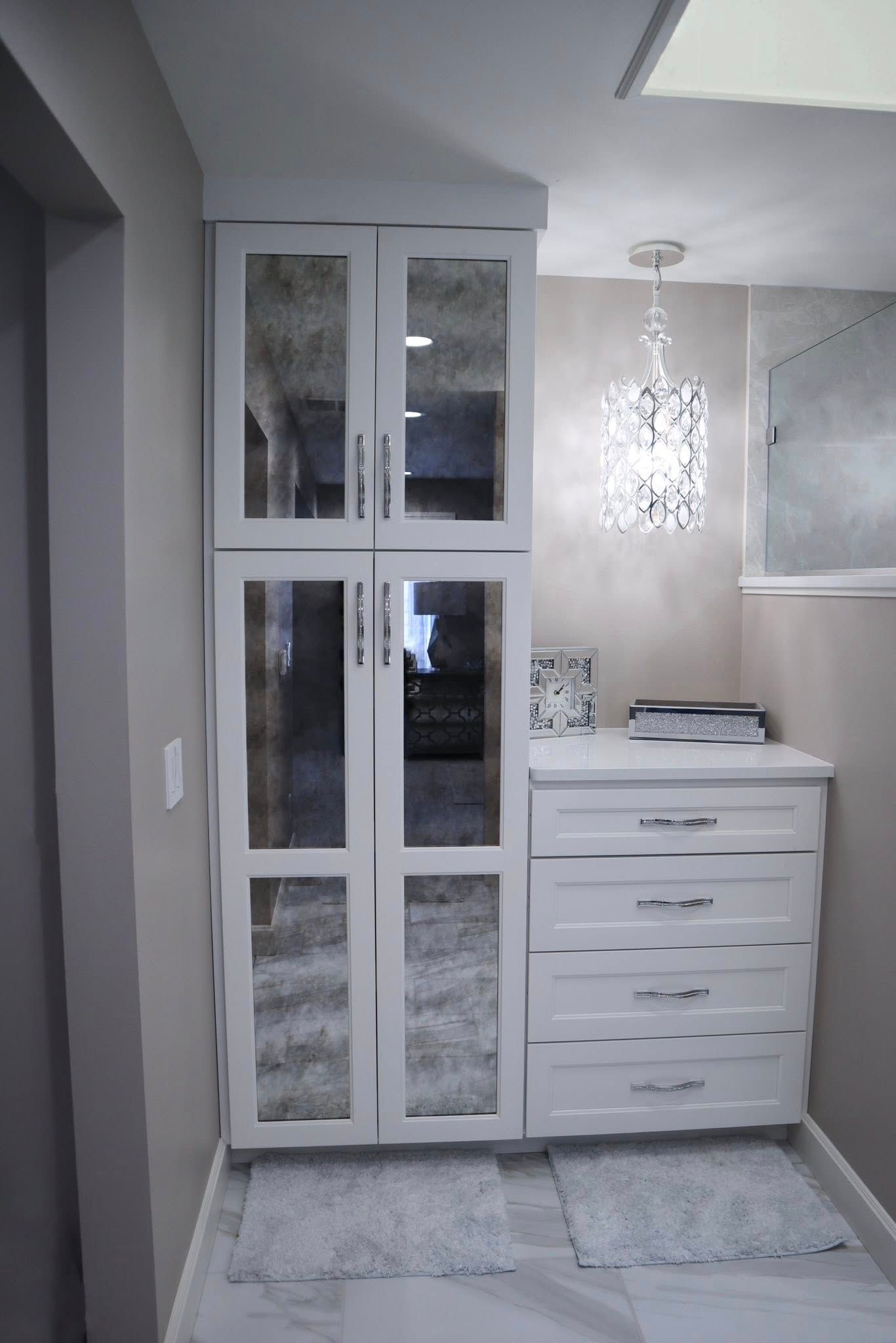 White tall mirror-fronted cabinet next to a white four-drawer chest in a hallway under a crystal pendant light.