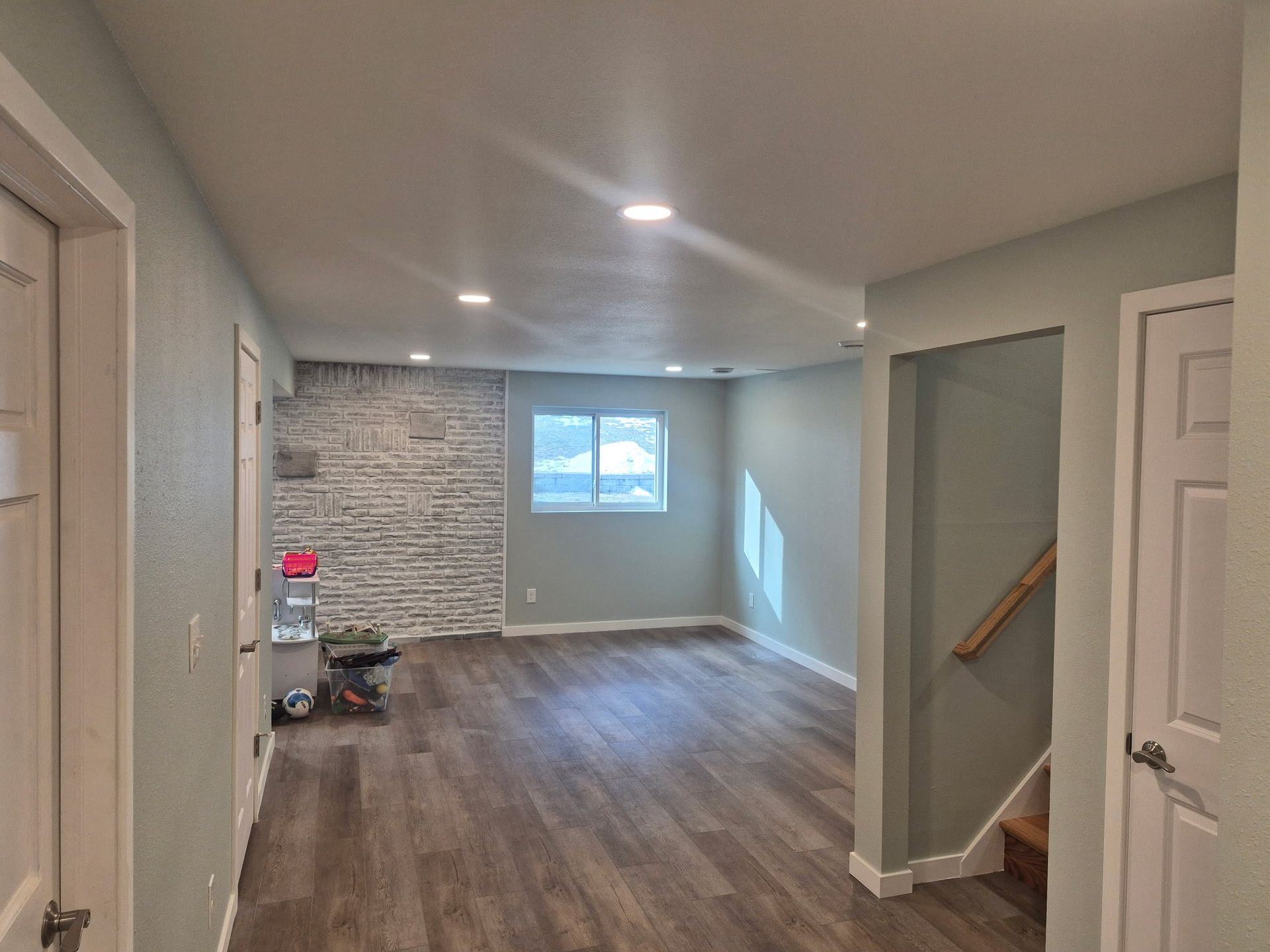 A view of a modern basement featuring wood-look floors, a white brick accent wall, light grey walls, and recessed lighting.